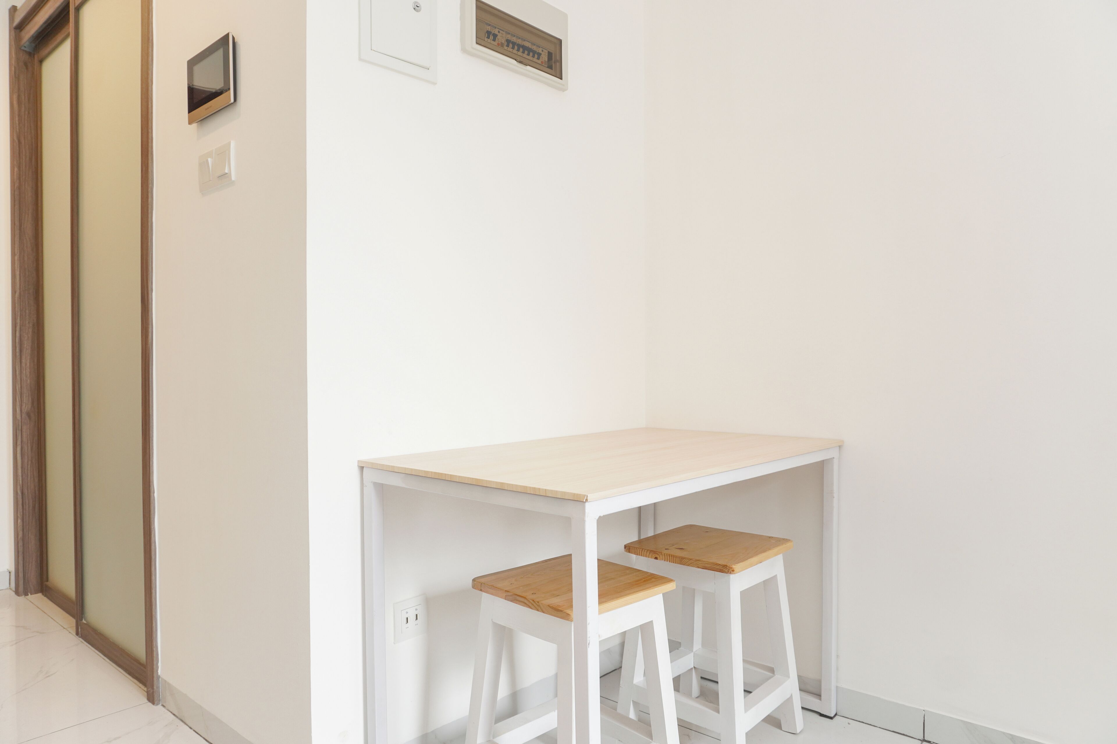 A small, white room with a wooden desk and two matching stools.