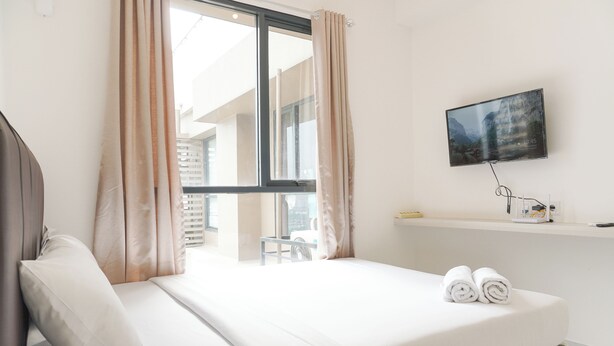 Ein Hotelzimmer mit einem Bett, einem an der Wand befestigten Fernseher, einem Fenster mit Vorhängen und Blick auf Gebäude.