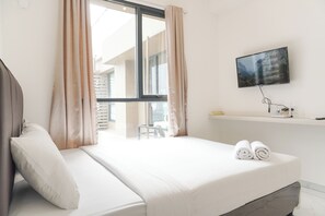 Quarto de hotel com cama, televisão na parede, janela com cortinas e vista para edifícios.