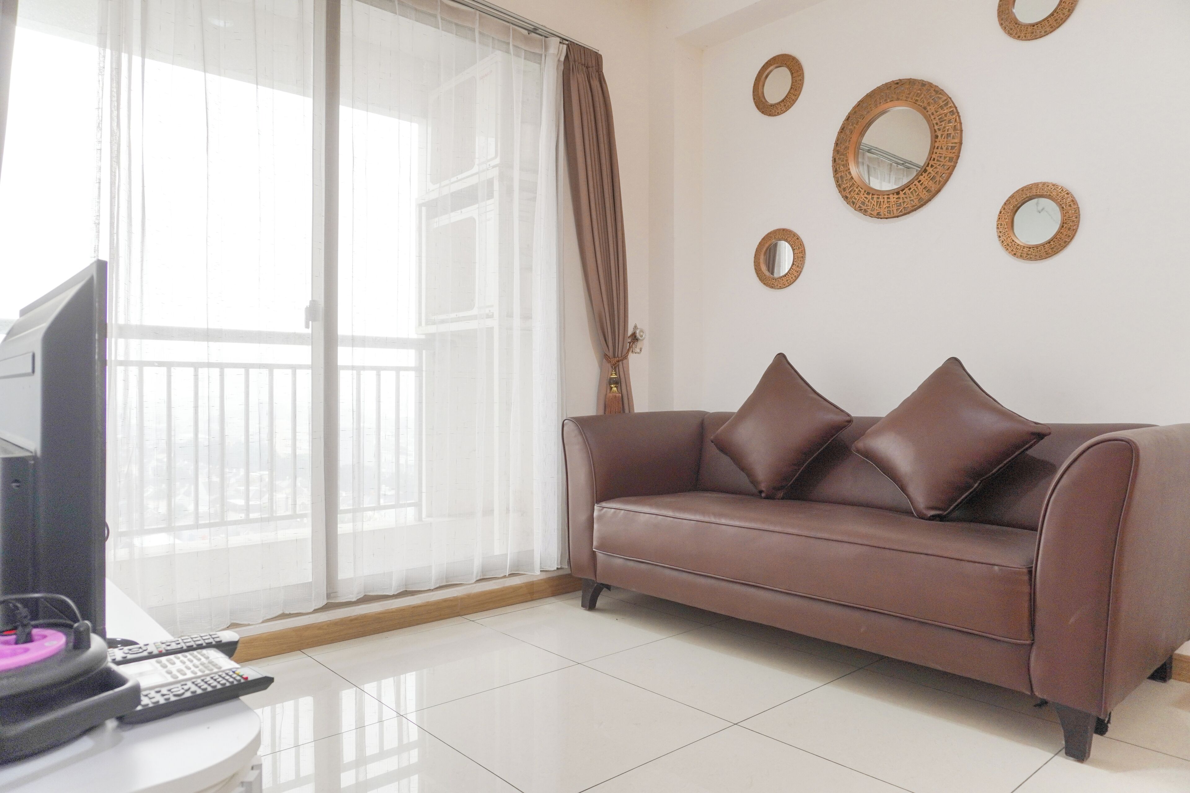 A living room with a brown leather sofa, a television, and a table with remote controls. There are curtains on the window, and decorative mirrors on the wall.
