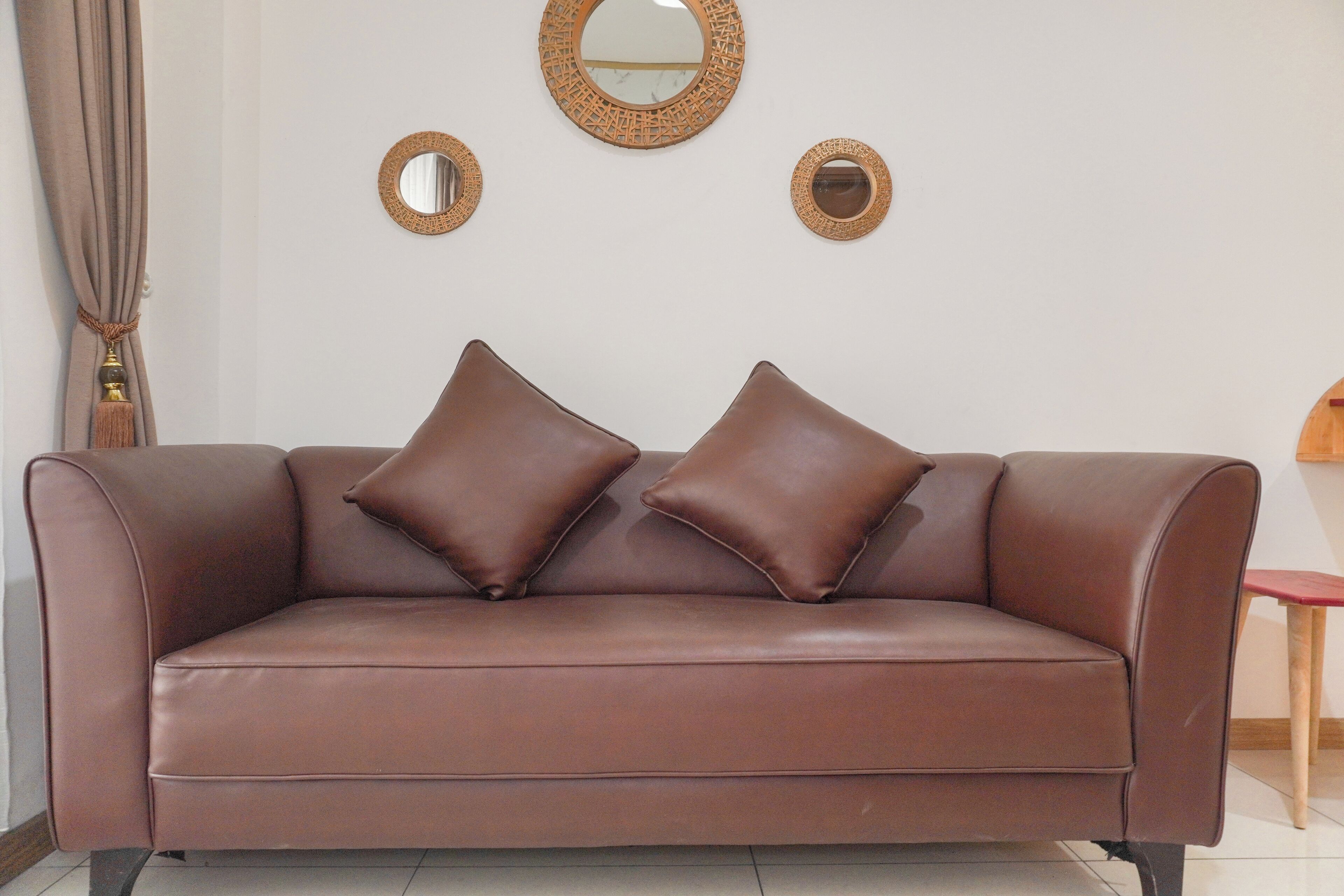 A brown leather sofa with two cushions, a round mirror, and a small wooden table in a room with white walls and a curtain.