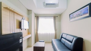 A modern living room with a black leather sofa, a flat-screen TV mounted on a wooden panel, a small table, and a window with curtains. - Homey And Minimalist 1Br Royal Sentul Park Apartment (Bogor)