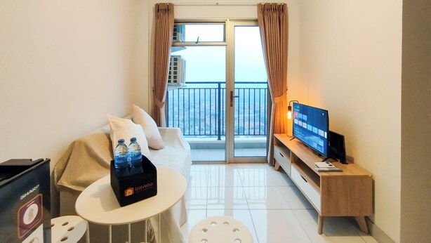 A small, well-lit room with a sofa, a small table with a bottle holder, a TV on a wooden stand, and a balcony with a view. - Modern 2Br At Mekarwangi Square Cibaduyut Apartment (Bandung)