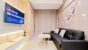 A modern living room with a flat-screen TV mounted on the wall, a black leather sofa, a small white table, and a decorative wall with cherry blossom patterns. - Modern Look And Cozy 1Br Branz Bsd City Apartment (Pagedangan)