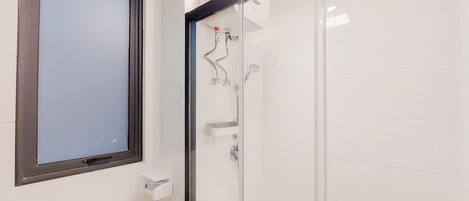 Un baño con inodoro, ducha con mampara de vidrio y ventana sobre el inodoro.