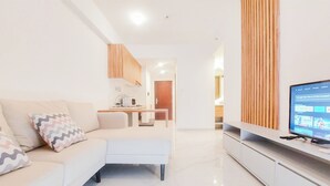 Uma sala de estar moderna com um sofá branco, uma televisão de tela plana e uma área de cozinha ao fundo.