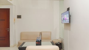 A room with a beige sofa, a small table with a water bottle, a television mounted on the wall, and a wooden door. - Best Deal And Restful 2Br Marina Ancol Apartment (Jakarta)