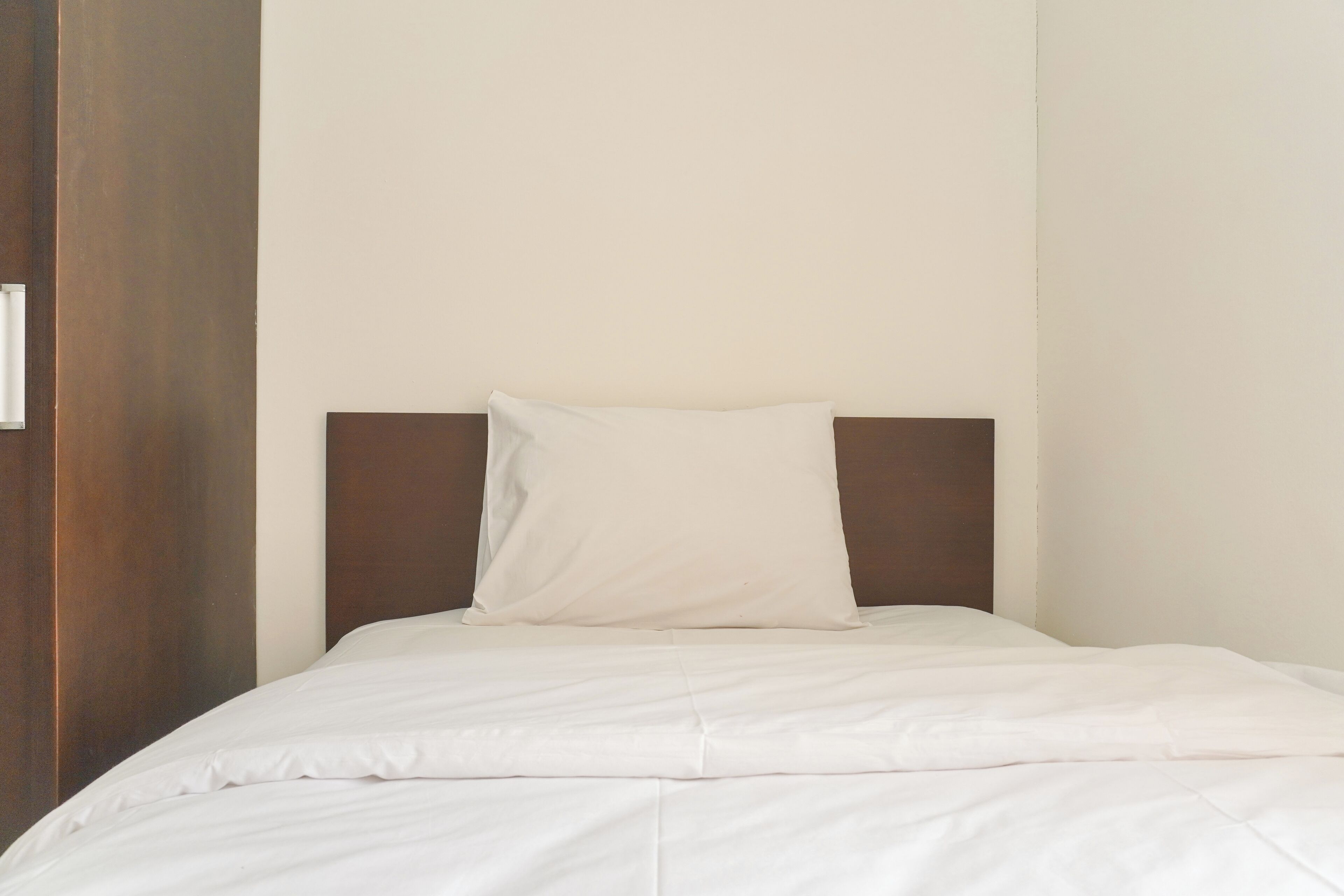 A neatly made bed with a white sheet and pillow in a room with a wooden headboard and a plain wall.