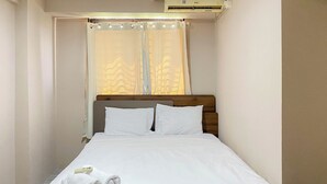 Un lit bien fait, avec des draps blancs et une serviette enroulée, une tête de lit en bois et une fenêtre ornée de rideaux légers.
