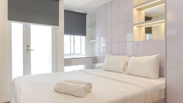 A modern hotel room with a large bed, a bedside table, a window with a view, and a bathroom door. - Compact Studio At Tokyo Riverside Apartment (Teluknaga)