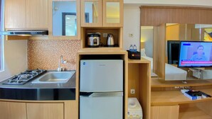 A compact kitchen with a stove, sink, and a built-in refrigerator. There is a television on a stand and a small shelf with a kettle and a box. - Cozy Living Studio At Kebagusan City Apartment (Jakarta)