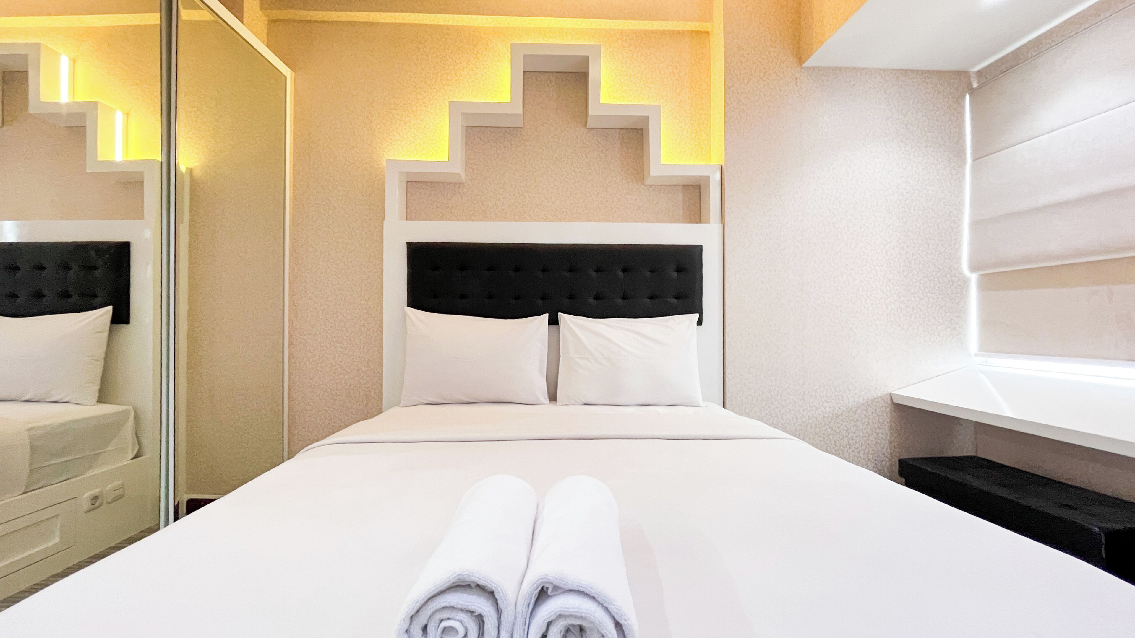 A hotel room with a neatly made bed, a black upholstered headboard, and a mirror reflecting the room.