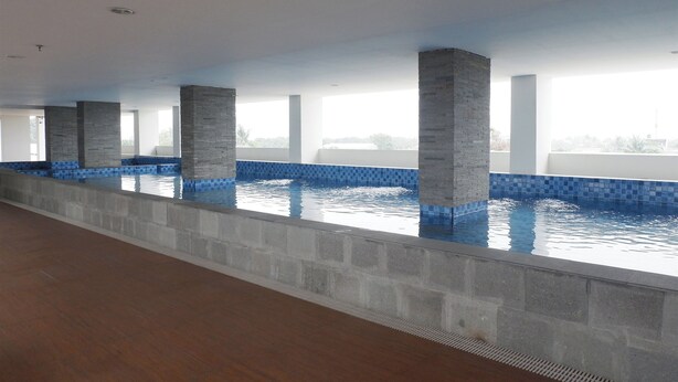 Une vaste piscine intérieure aux bords recouverts de carreaux bleus, soutenue par des piliers gris et entourée d’un haut plafond doté d’un éclairage encastré.