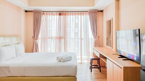 A hotel room with a bed, a desk with a TV, and large windows with curtains. - Well Furnished And Cozy Studio Casa De Parco Apartment (Pagedangan)