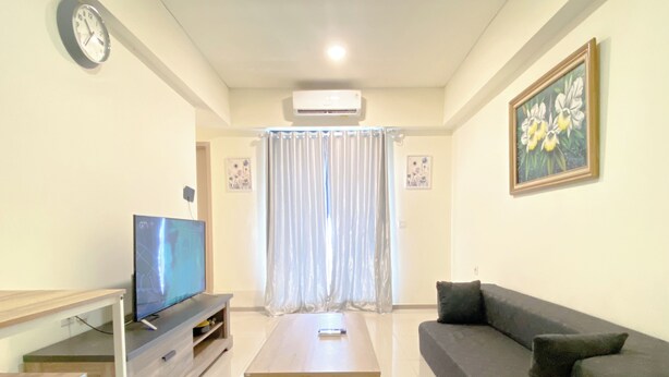 A living room with a flat-screen TV, a wooden coffee table, a dark sofa, and a painting on the wall. - Comfortable Living 2Br At Meikarta Apartment (Cikarang)