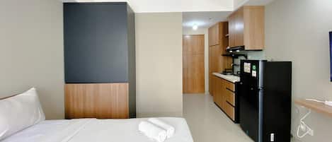 A hotel room with a bed, a wardrobe, a kitchenette with a refrigerator, and a wooden door leading to another room.