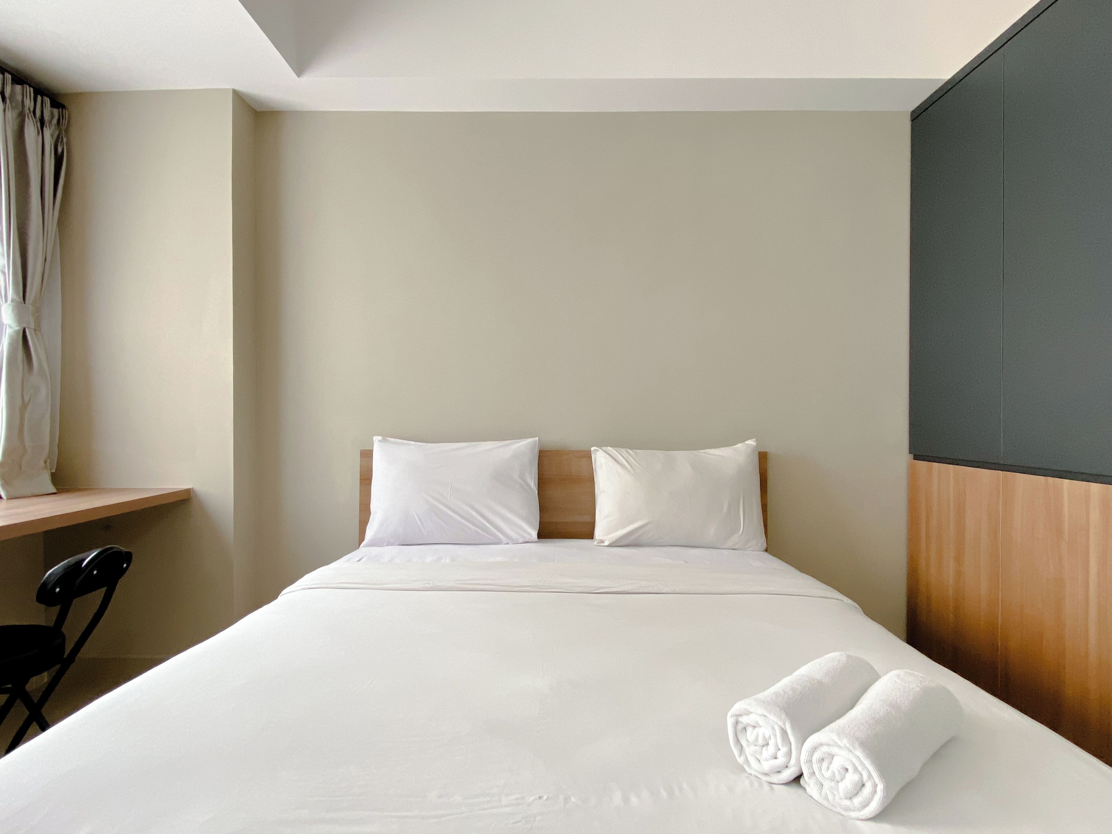 Une chambre d’hôtel avec un lit bien fait, deux oreillers, une tête de lit en bois, une table de chevet en bois, une chaise noire et une fenêtre avec des rideaux.
