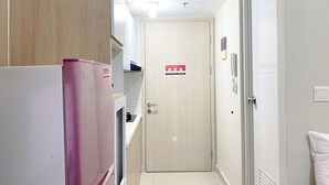 A narrow hallway with a door at the end, a refrigerator on the left, and a white wall with a light switch and a phone on the right.