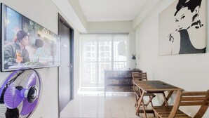 A room with a TV mounted on the wall, a purple fan, a wooden table with chairs, and a large window with a view of buildings. - Modern Look 2Br At Serpong Greenview Apartment (South Tangerang)