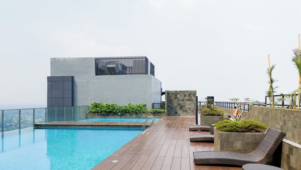 A rooftop infinity pool with a wooden deck, lounge chairs, and a modern building with a glass railing.