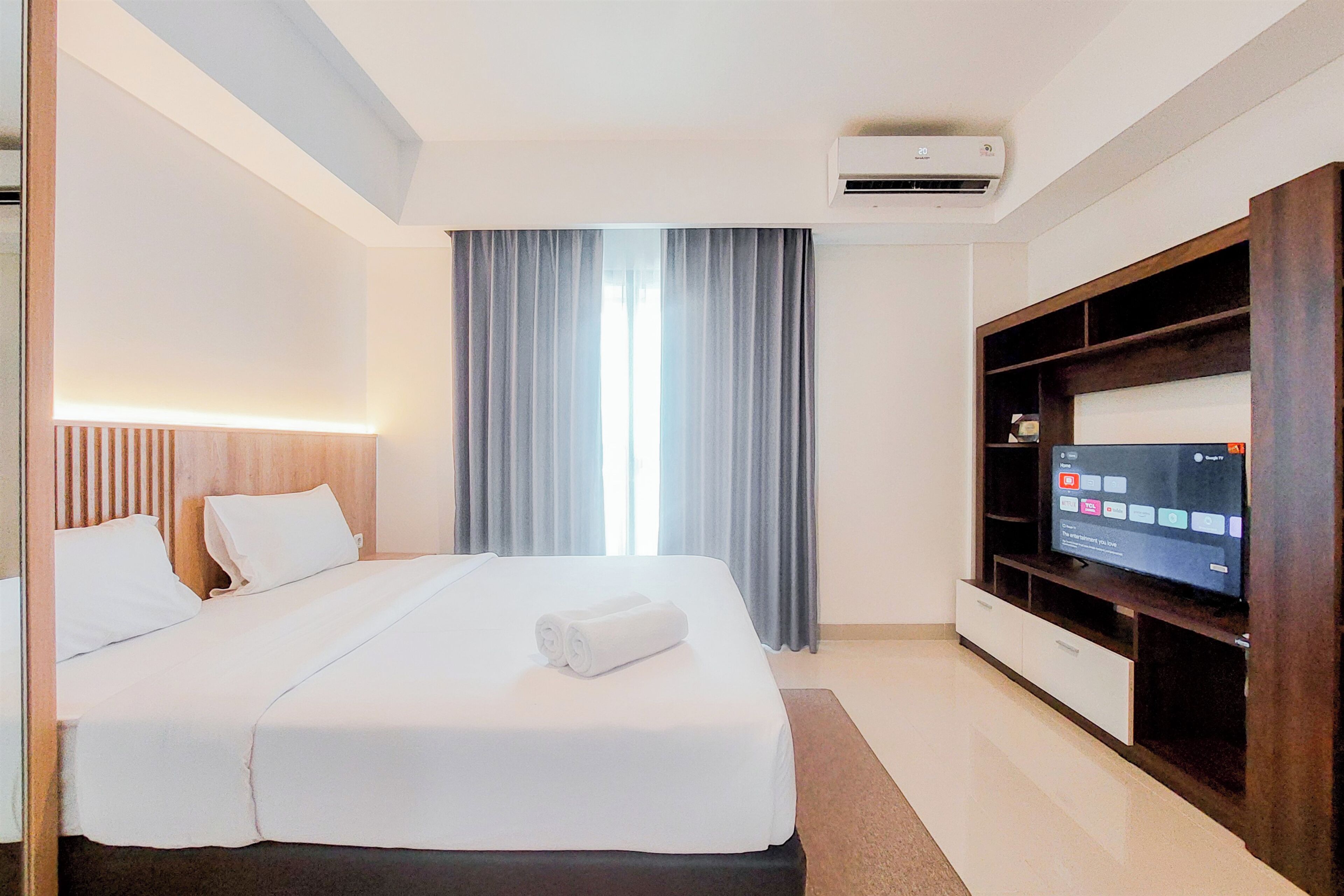 A hotel room with a bed, a TV, a window with curtains, and an air conditioning unit.
