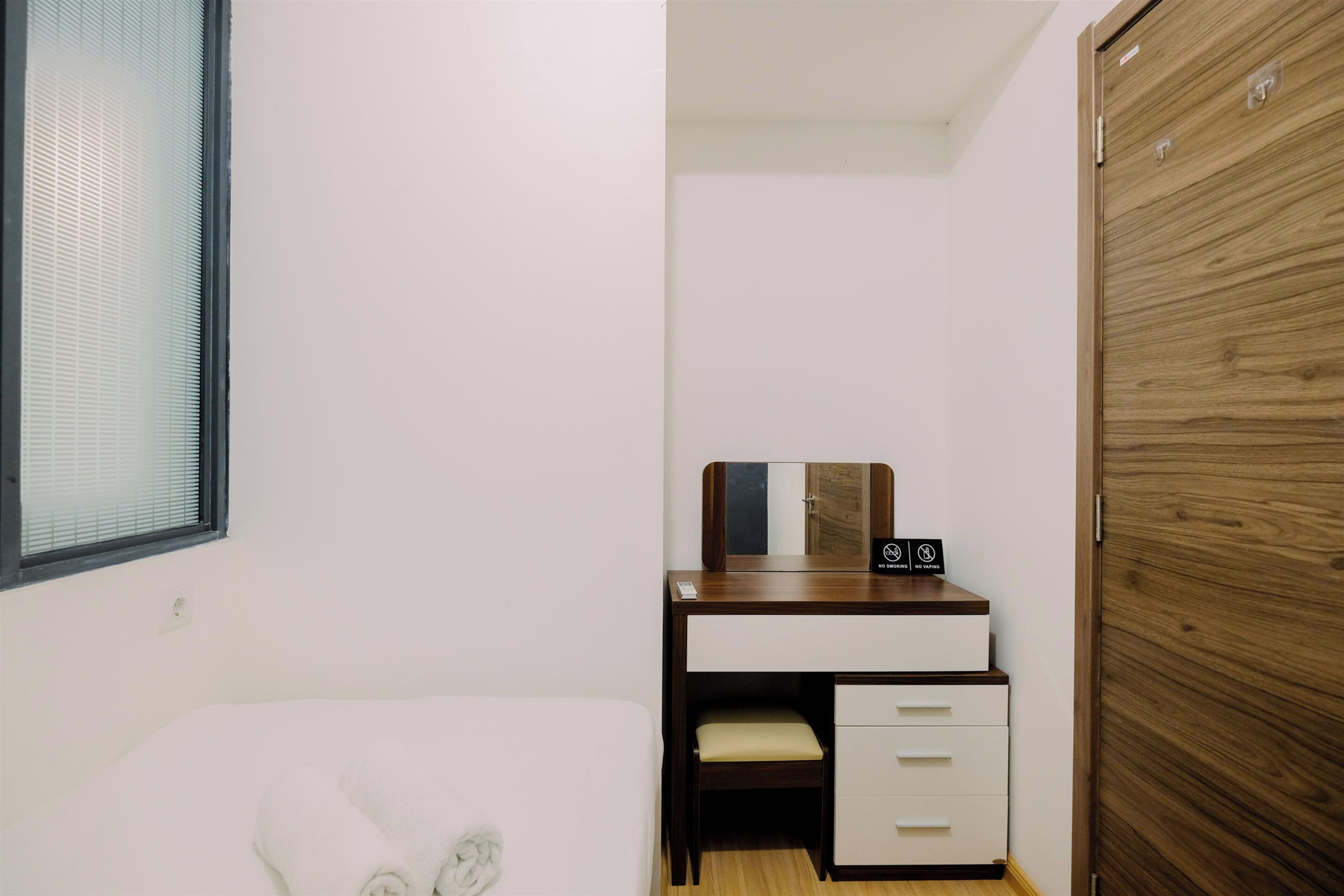 A bedroom with a bed, a wooden wardrobe, a desk with a mirror, and a window with blinds.