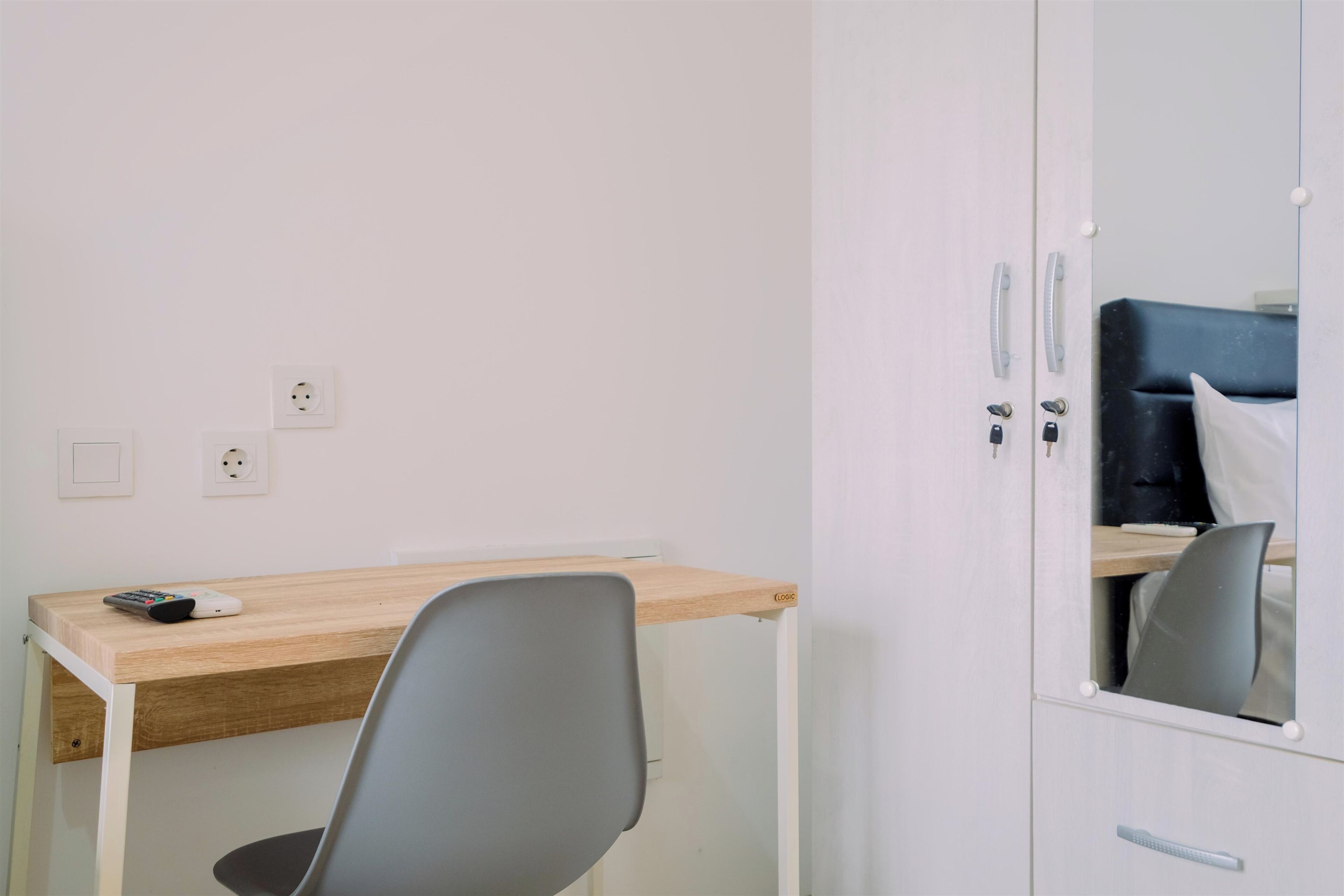 A minimalist room with a wooden desk, a grey chair, and a wardrobe with a mirror.
