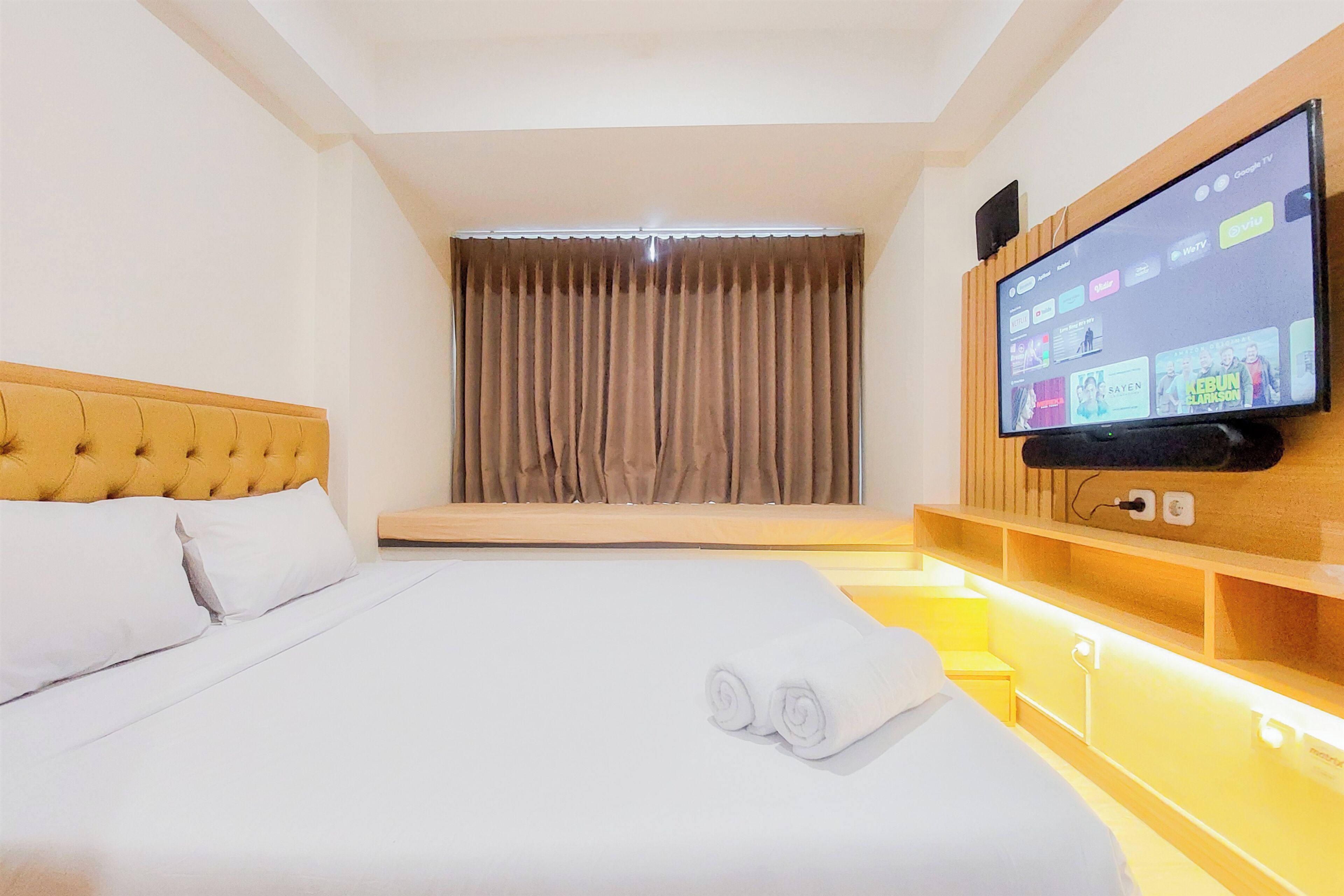 Une chambre d’hôtel avec un grand lit, un téléviseur à écran plat fixé au mur, une fenêtre avec des rideaux et une tête de lit en bois.