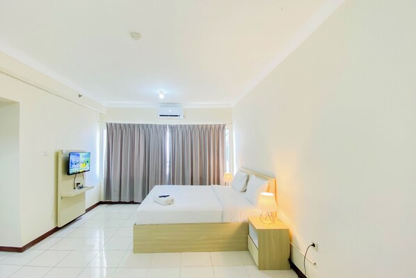 A hotel room with a bed, a TV on a stand, a bedside table with a lamp, and a window with curtains.