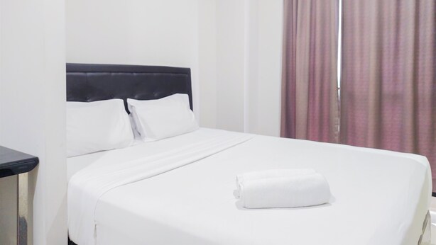 Una cama bien tendida con sábanas blancas y una toalla enrollada, un cabecero negro y una ventana con cortinas moradas.