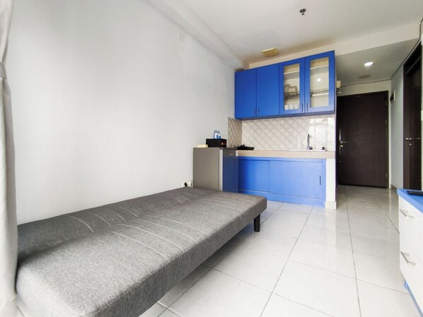 A compact room with a grey sofa, a small kitchenette with blue cabinets, and a white tiled floor. - Spacious And Cozy 2Br Apartment Mekarwangi Square Cibaduyut (Bandung)