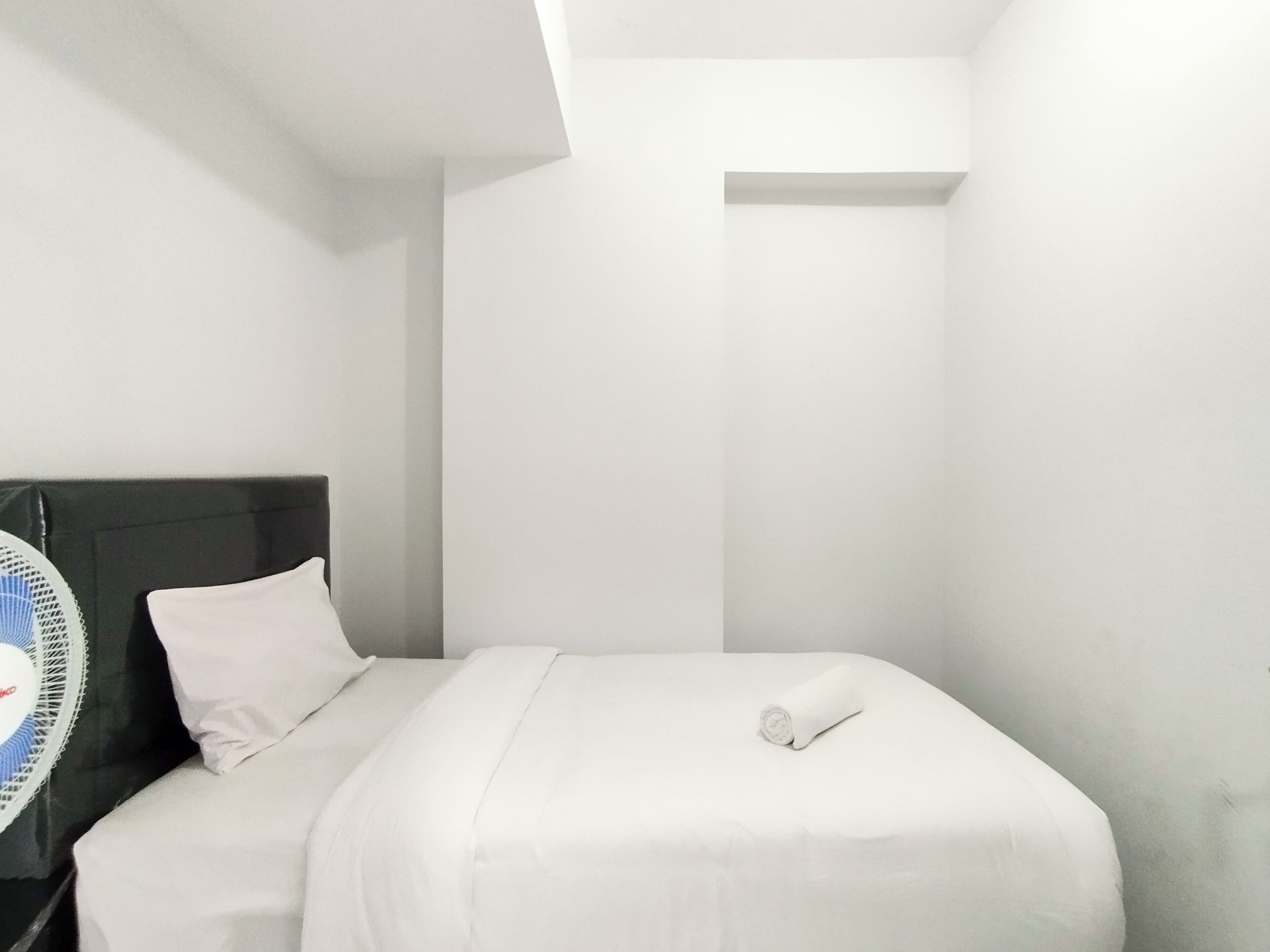 Un petit lit bien fait, avec du linge de lit blanc et une serviette enroulée, une table de chevet avec un ventilateur et un mur blanc sobre.