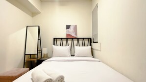 A hotel room with a neatly made bed, a bedside table with a lamp, a mirror, and a painting on the wall. - Clean And Homey 2Br At Meikarta Apartment (Cikarang)