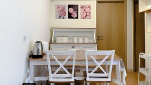 A small kitchen with a wooden table, two white chairs, and a white cabinet with a wooden countertop. There are three framed pictures of pink flowers on the wall above the cabinet. - Clean And Homey 2Br At Meikarta Apartment (Cikarang)