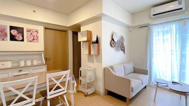 A modern living room with a sofa, a dining table with chairs, a small side table, and a wall-mounted air conditioner. - Clean And Homey 2Br At Meikarta Apartment (Cikarang)