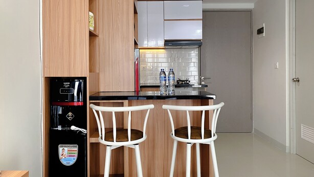 A compact kitchen with wooden cabinets, a black refrigerator, and two white bar stools. - Well Furnished And Spacious 1Br Vasanta Innopark Apartment (Cikarang)