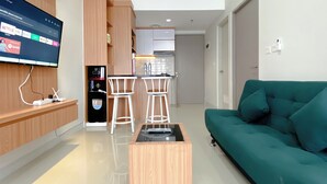 Ein modernes Wohnzimmer mit einem salbeengrünfarbenen Sofa, einem Holztisch und einem an der Wand montierten Fernseher. Im Hintergrund sind zwei weiße Barhocker und eine Küchenzeile zu sehen.