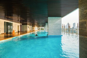 An indoor swimming pool with clear blue water, a city skyline view through large windows, and a modern architectural design with a sloped ceiling. - Comfort And Modern Look Studio Menteng Park Apartment (Jakarta)