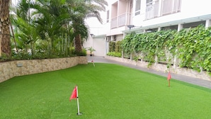 A well-maintained artificial grass area with a red flag marker, surrounded by stone walls and lush greenery. - Simply Look Studio At Mustika Golf Residence (No Kitchen) Apartment (Cikarang)