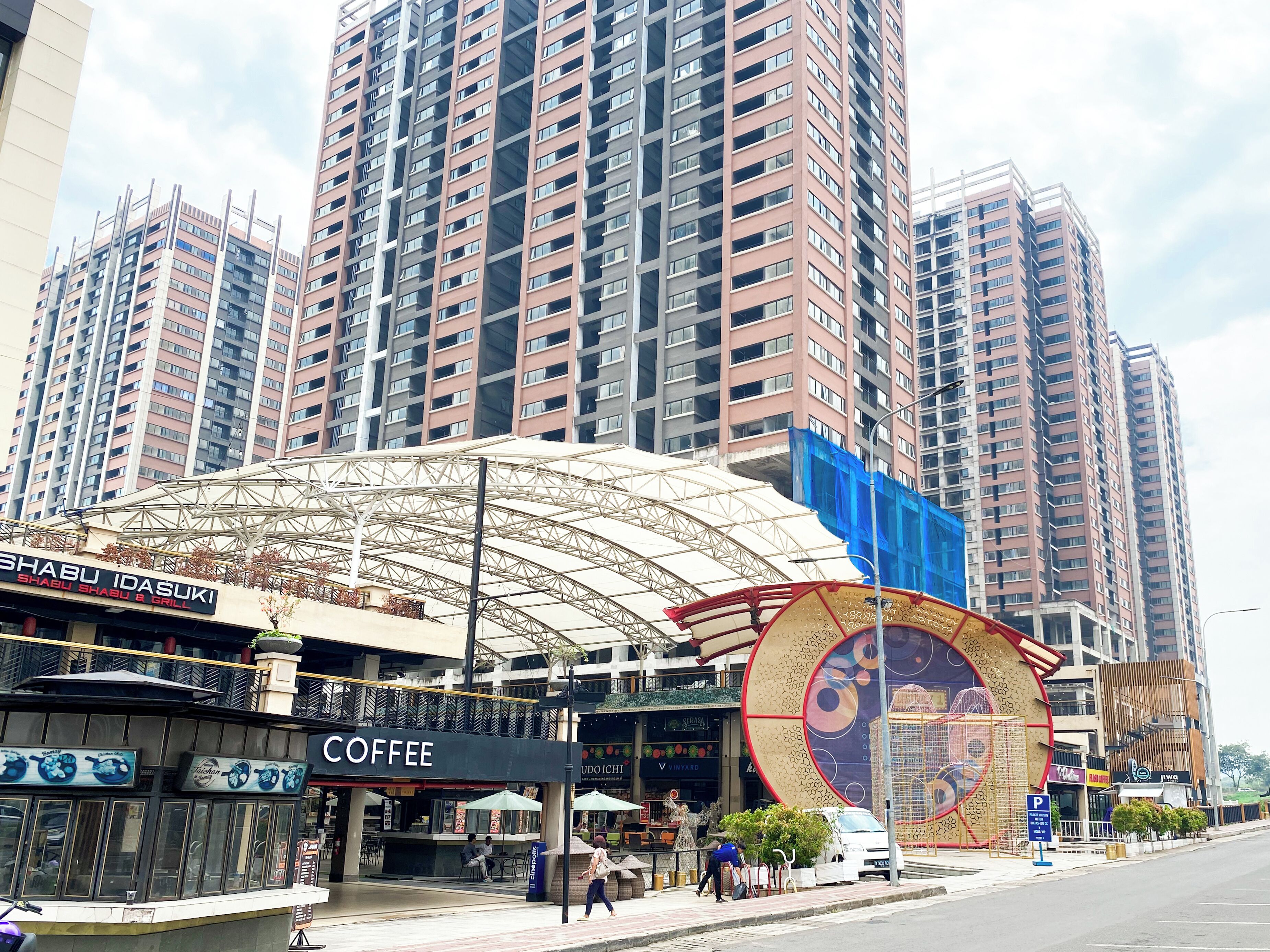 A modern urban street with high-rise buildings, a covered outdoor seating area, and various commercial establishments including a coffee shop.