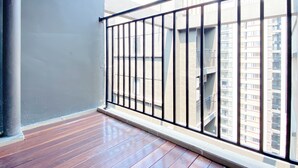 A balcony with a wooden floor and metal railing, overlooking a cityscape with high-rise buildings.