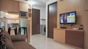 A modern living room with a sofa, a television, and a kitchen area with a microwave and washing machine. - Good Deal And Comfort Stay 2Br Transpark Cibubur Apartment (Depok)