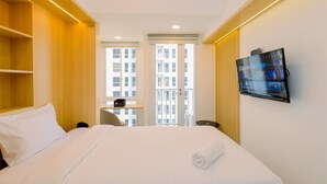 A hotel room with a bed, a TV mounted on the wall, a desk with a chair, and a view of buildings through the window. - Simply Look And Cozy Studio Tokyo Riverside Pik 2 Apartment (Teluknaga)
