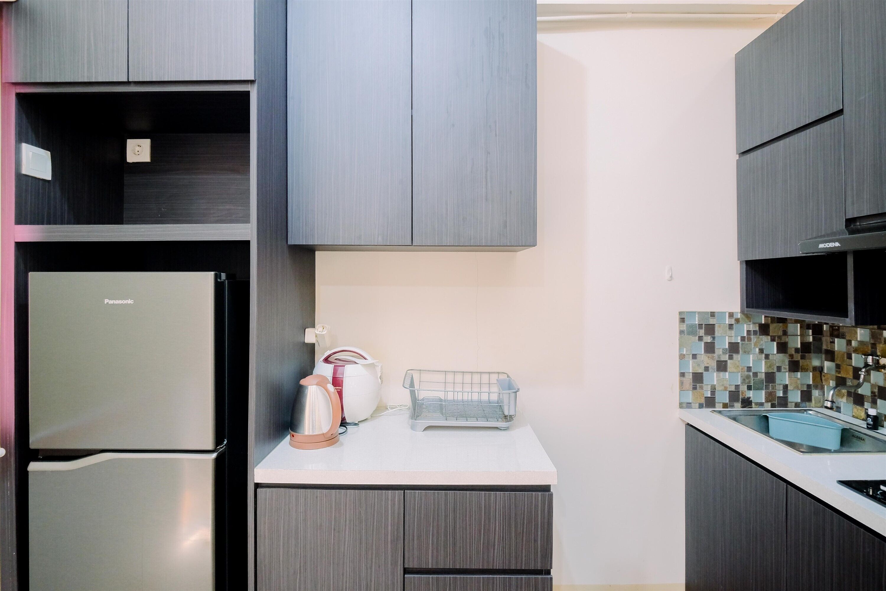 Eine kompakte Küche mit grauen Schränken, einer weißen Arbeitsplatte, einem Panasonic-Kühlschrank, einem Wasserkocher und einem kleinen Spülbecken.