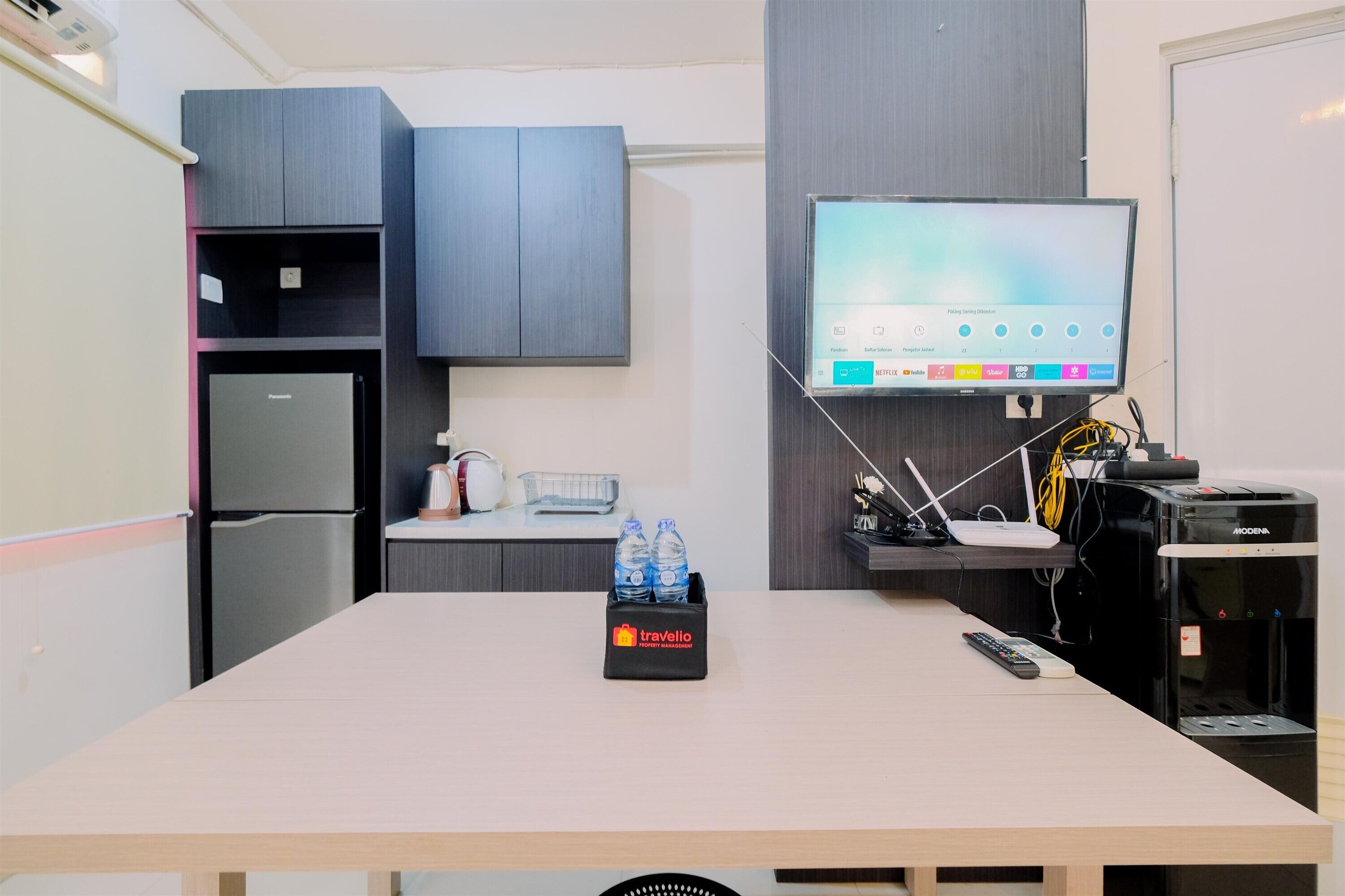 Une chambre moderne équipée d’un téléviseur à écran plat fixé au mur, d’un réfrigérateur, d’un distributeur d’eau et d’une trousse de toilette posée sur la table.
