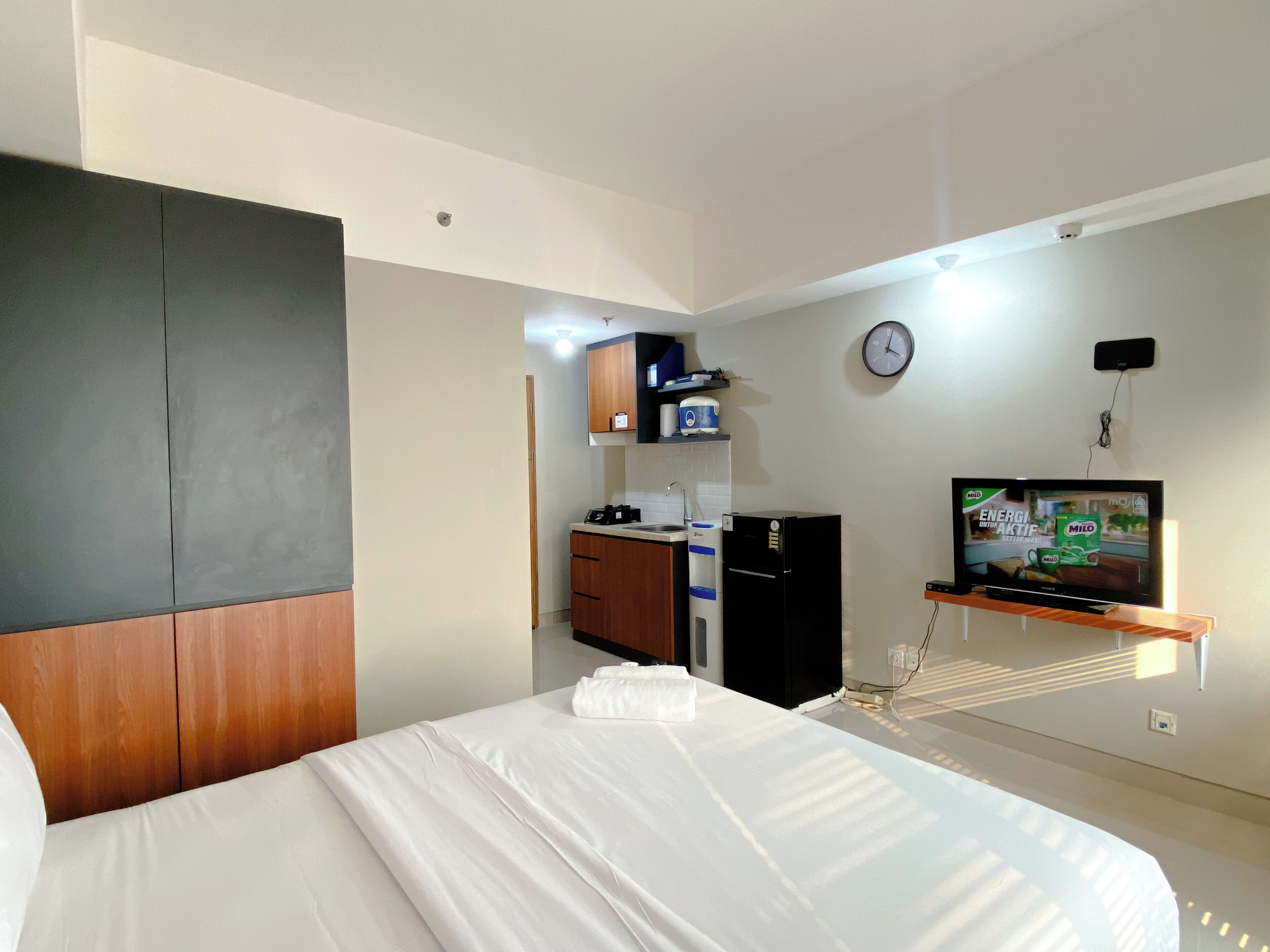 Habitación de hotel con cama, televisor, kitchenette y un reloj en la pared.