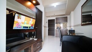 A modern living room with a flat-screen TV mounted on a wooden cabinet, a dining area with a table and chairs, and a kitchen with dark wooden cabinets and a tiled backsplash. - Best Price And Homey 1Br At Tamansari Prospero Apartment (Sidoarjo)
