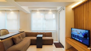 A modern living room with a sectional sofa, a flat-screen TV mounted on a wooden wall, and a large window with blinds. - Spacious Classic Studio At Jarrdin Cihampelas Apartment (Bandung)