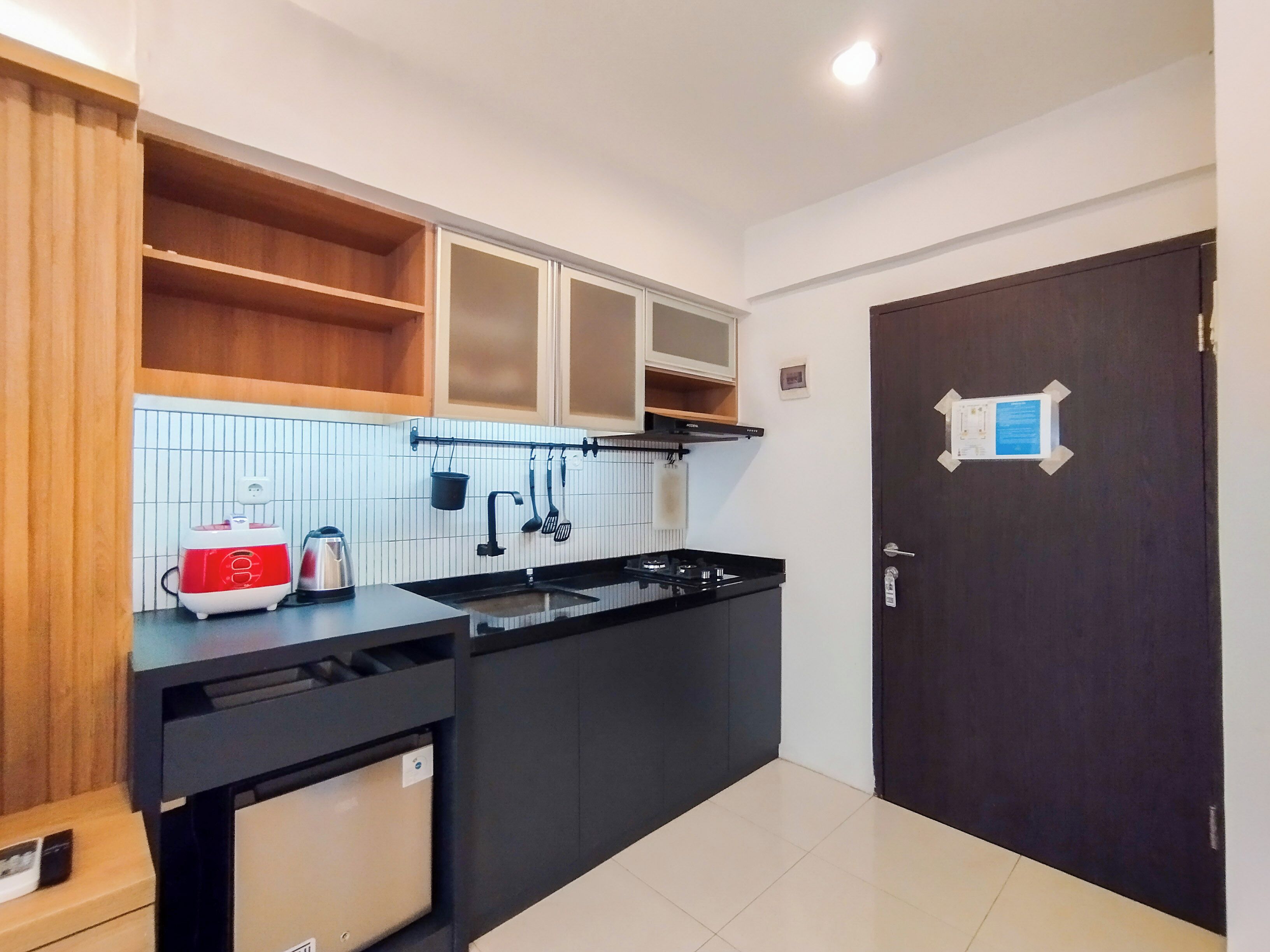 A compact kitchen with a black countertop, a red and white coffee maker, a kettle, and a stove. There is a wooden shelf above the countertop and a dark door with a sign on it.
