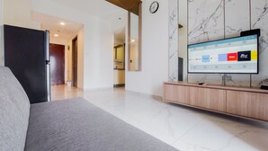 A modern living room with a grey sofa, a wooden TV stand, and a large flat-screen TV displaying a streaming service interface. - Comfort Living 2Br At Sky House Bsd Apartment (Pagedangan)